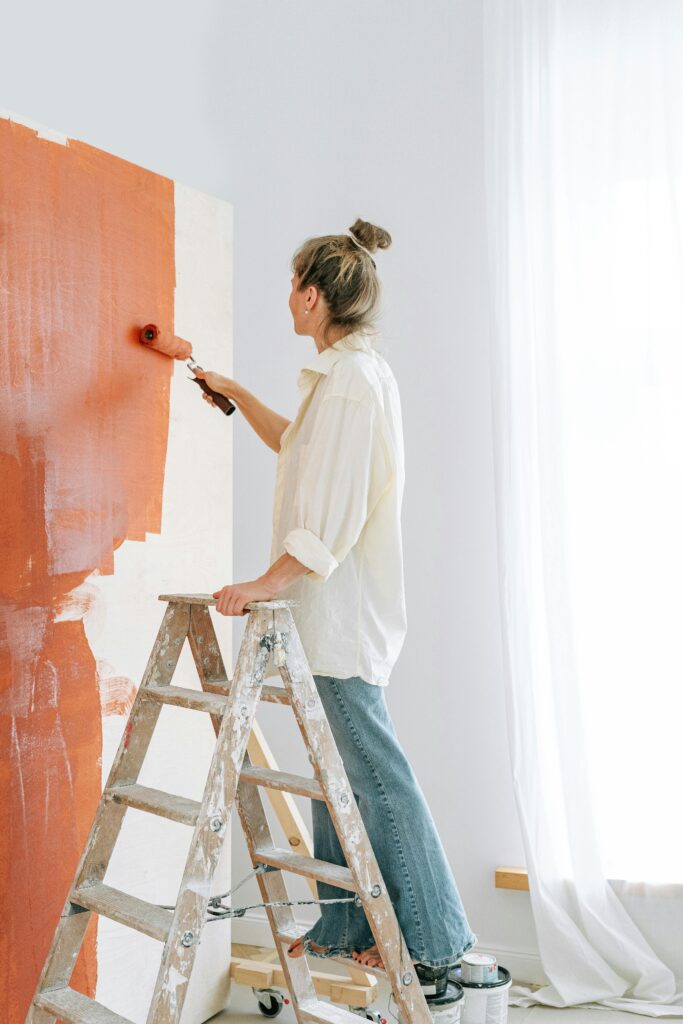 pexels-photo-5799049-5799049 An adult woman painting a wall with red paint using a roller indoors, standing on a ladder.