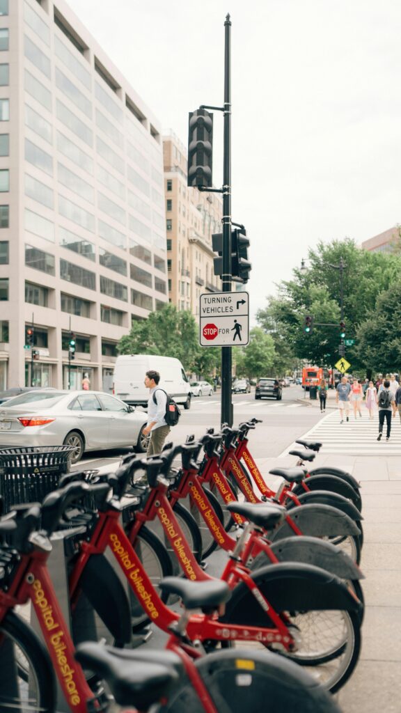 pexels-photo-26546821-26546821 Red city bikes lined up on an urban street with people walking and cars passing by. Ideal for city life themes.